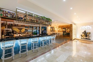A bar area with multiple shelves of liquor bottles, hanging wine glasses, and white wicker bar stools. - Hotel Andes Plaza (Bogotá)