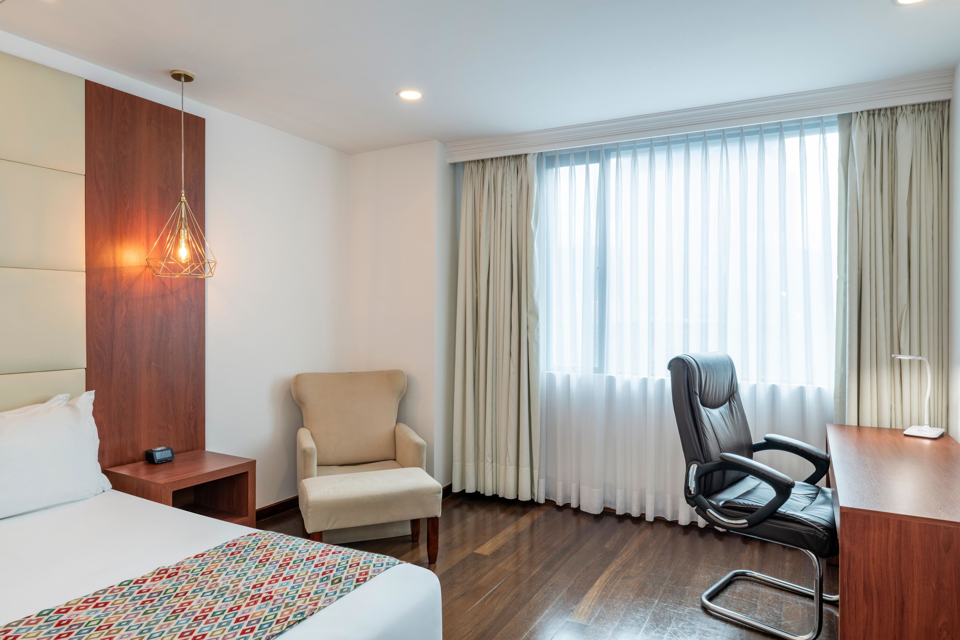 A hotel room with a bed, a desk, a chair, a desk lamp, a wall-mounted light fixture, and a window with curtains.