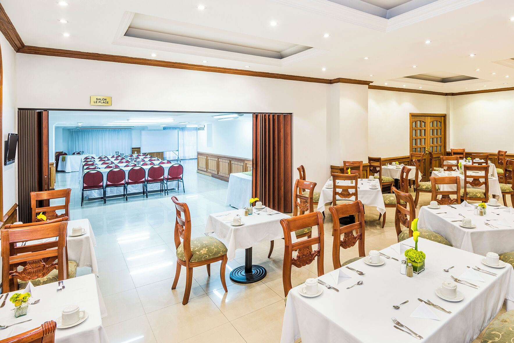 A hotel dining area with multiple tables and chairs, a conference room in the background, and a sign indicating the location of the salon.