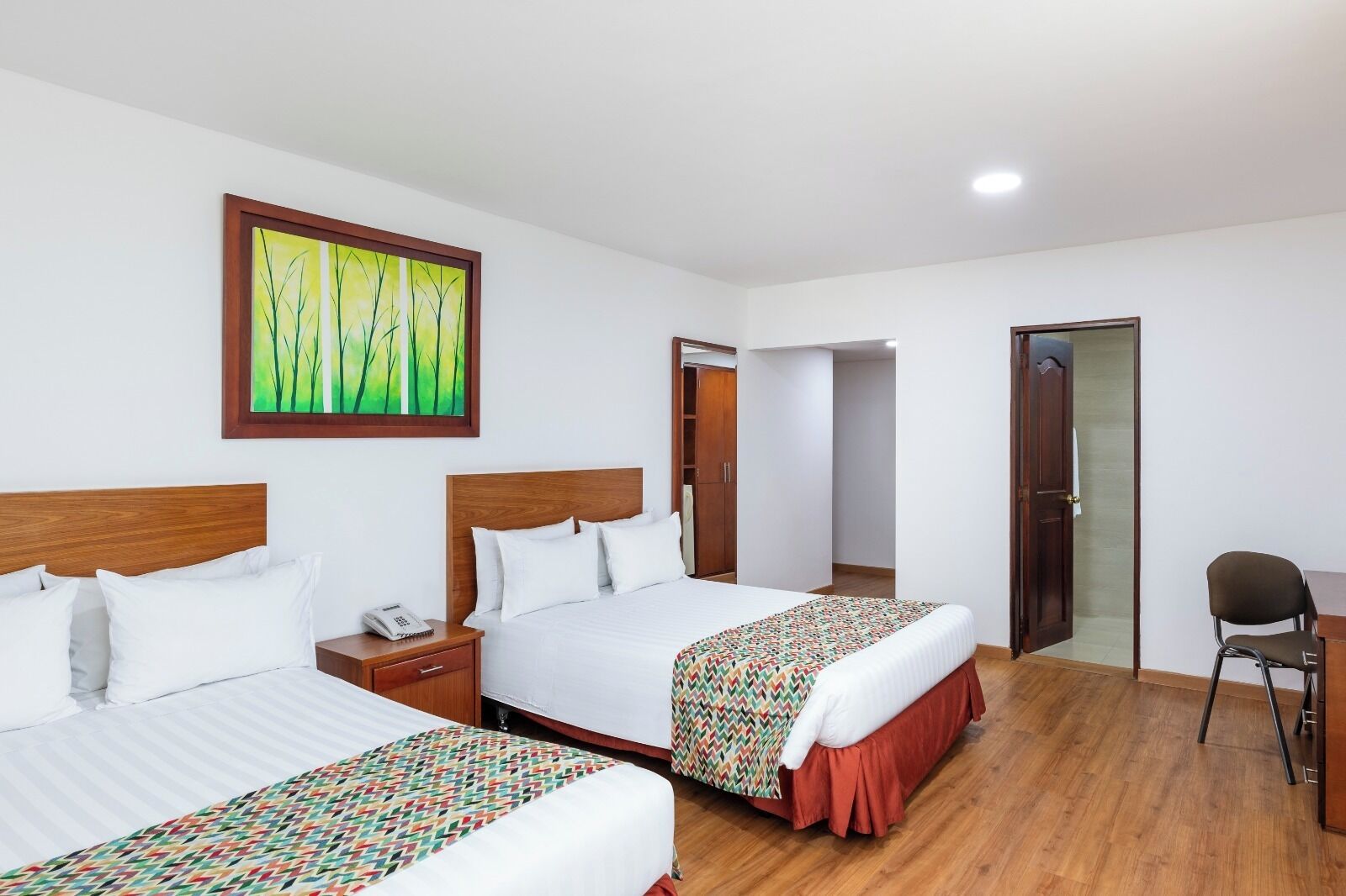 A hotel room with two beds, a wooden headboard, a bedside table with a phone, a colorful blanket, and a painting on the wall.