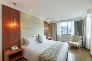 Habitación de hotel con una cama grande, una mesita de noche con teléfono, una silla, vista a la ciudad y una manta decorativa.