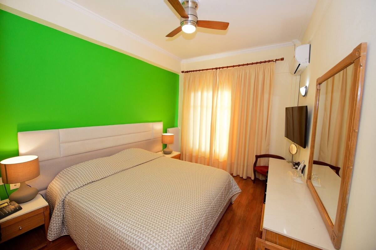 A bedroom with a green wall, a bed with a patterned bedspread, a nightstand with a lamp, a dresser with a mirror, and a ceiling fan.
