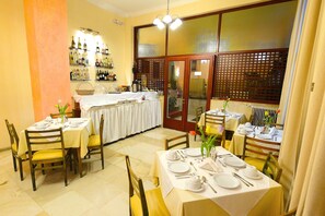A dining area with tables set for four, each with white tablecloths and silverware. There are chairs with yellow cushions, and a large window with a wooden lattice design.