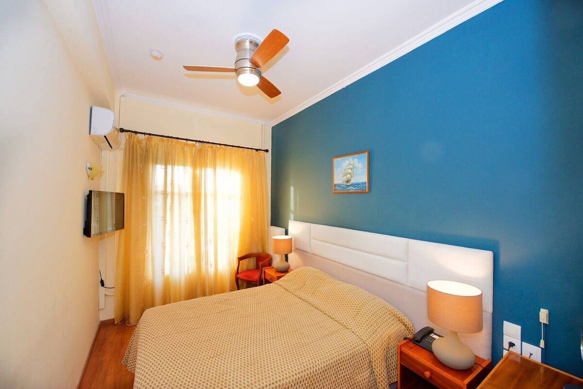 A bedroom with a bed, a nightstand, a lamp, a ceiling fan, a window with curtains, and a wall-mounted TV.