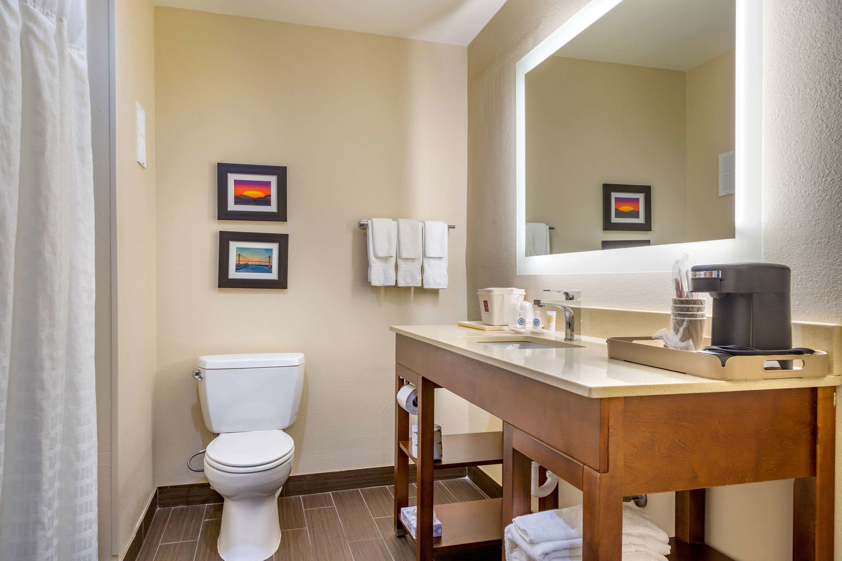 Un bagno con il water, un lavandino e uno specchio. Asciugamani appesi al muro e una macchina per il caffè sul ripiano.