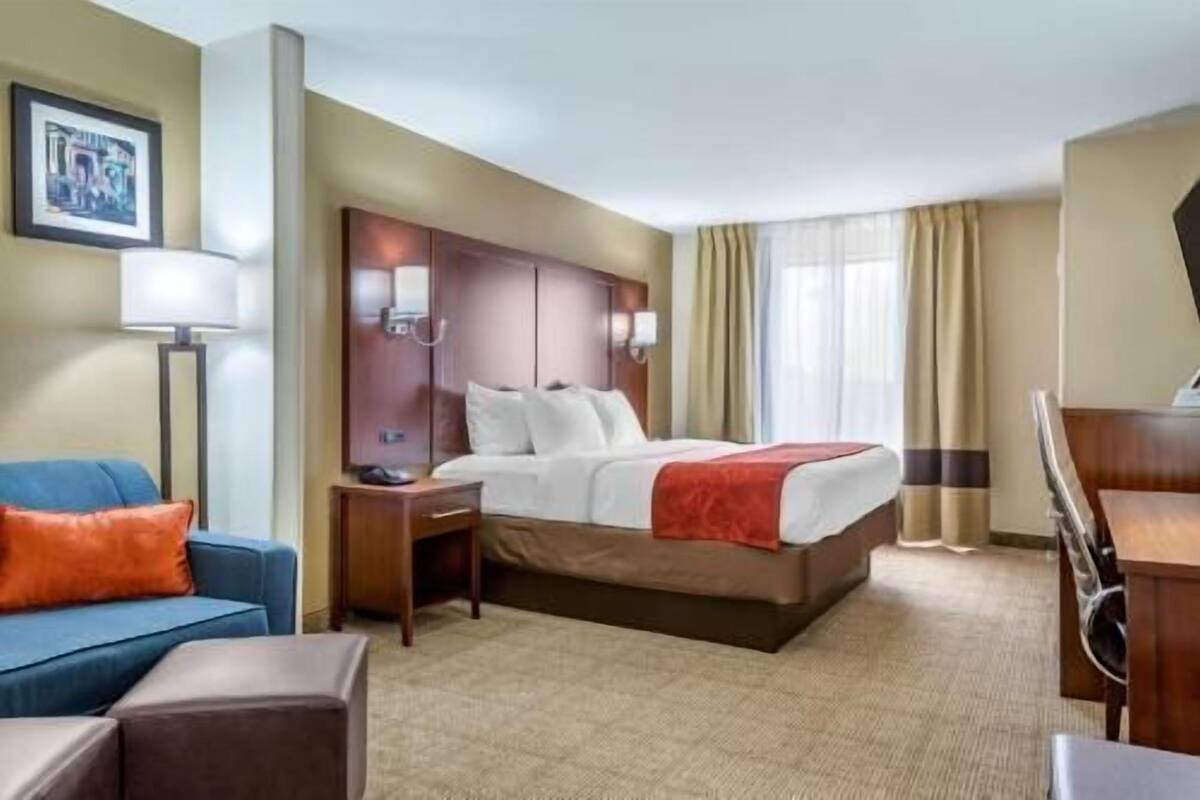A hotel room with a bed, a desk, a chair, a lamp, a sofa, and a window with curtains.