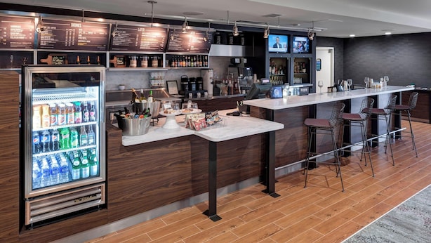 Un espace bar moderne doté d’un présentoir réfrigéré pour les boissons, d’un long comptoir avec des tabourets et d’un panneau affichant le menu au-dessus.