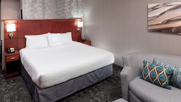 Ein Hotelzimmer mit einem großen Bett, zwei Nachttischen mit Lampen, einem grauen Sofa mit gemusterten Kissen und einer Wand mit abstrakter Kunst.