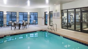 Une piscine donnant sur une pièce baignée de lumière, où se trouvent une table et des chaises, ainsi qu’un espace dédié à la musculation avec des tapis de course.