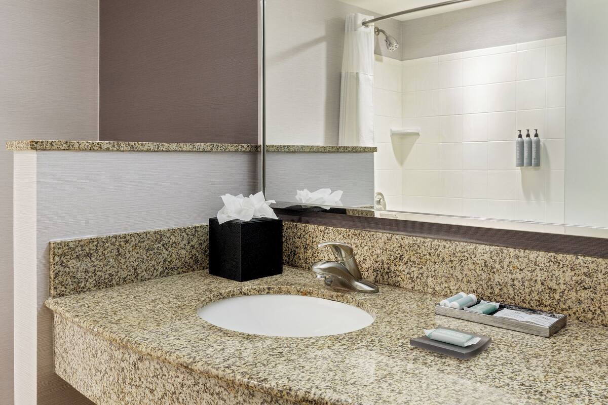 A bathroom with a granite countertop, a white sink, a mirror, a black tissue box, and toiletries.