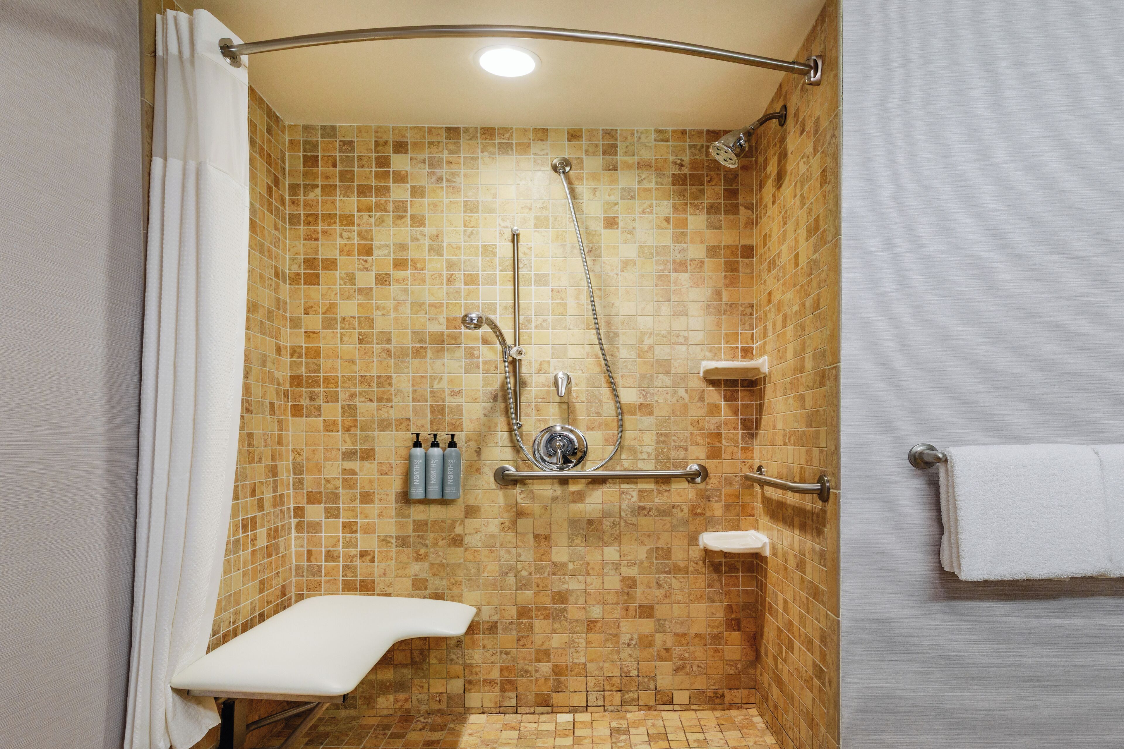A shower with a tiled wall, a handheld showerhead, a wall-mounted faucet, and a small shelf with toiletries.