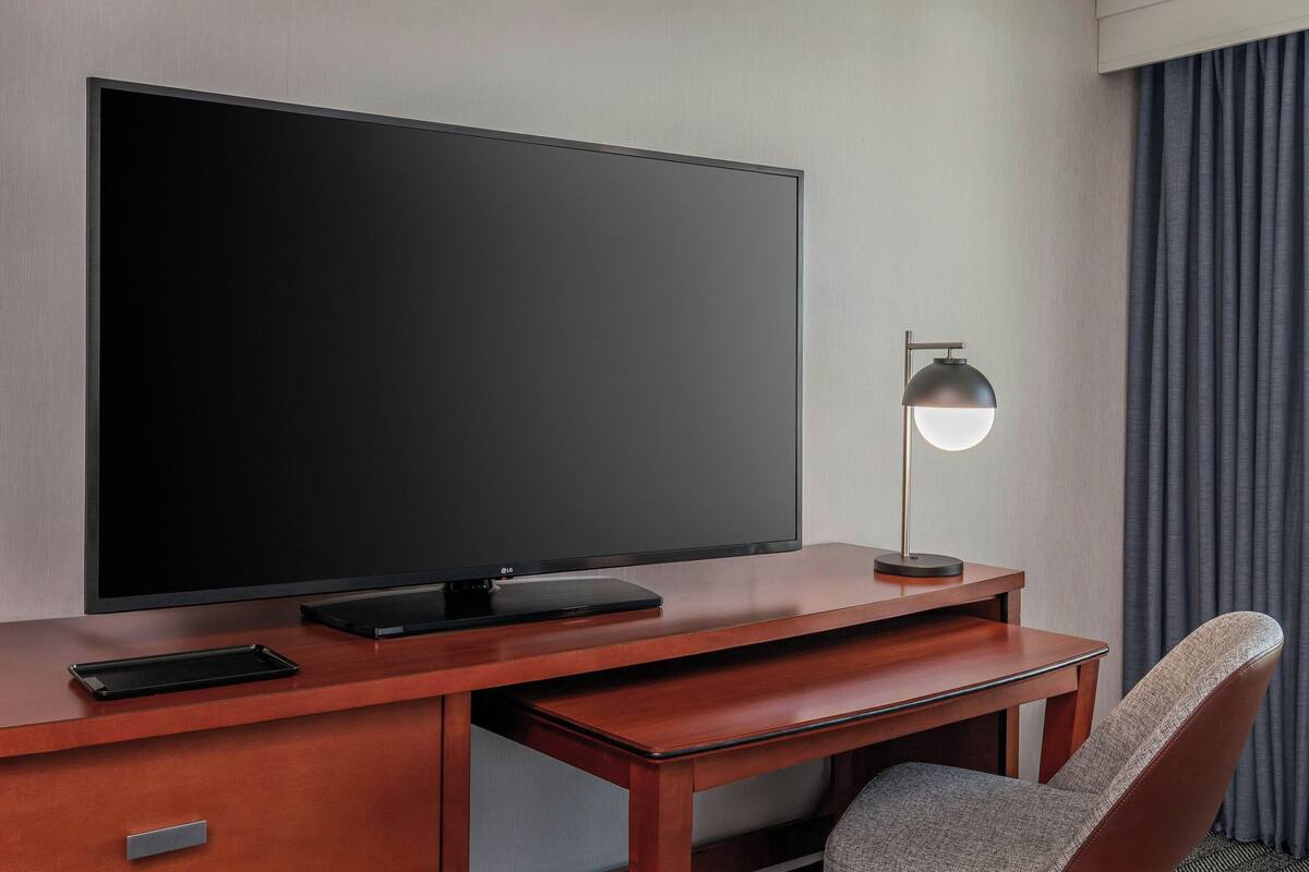 A hotel room with a large flat-screen TV on a wooden desk, a lamp, a chair, and a small drawer.