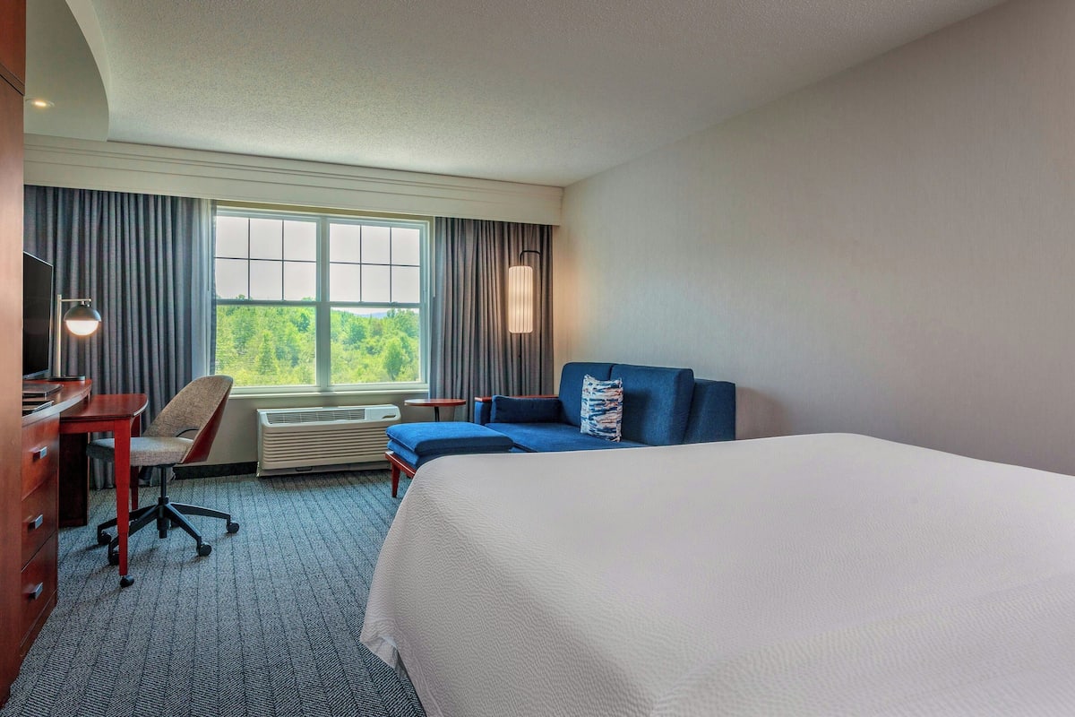 A hotel room with a large bed, a desk with a chair, a sofa, a standing lamp, and a large window with curtains.