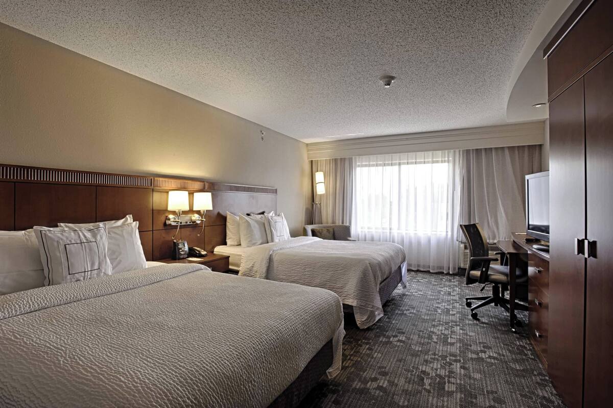 A hotel room with two beds, a desk, a chair, a TV, and a window with curtains.