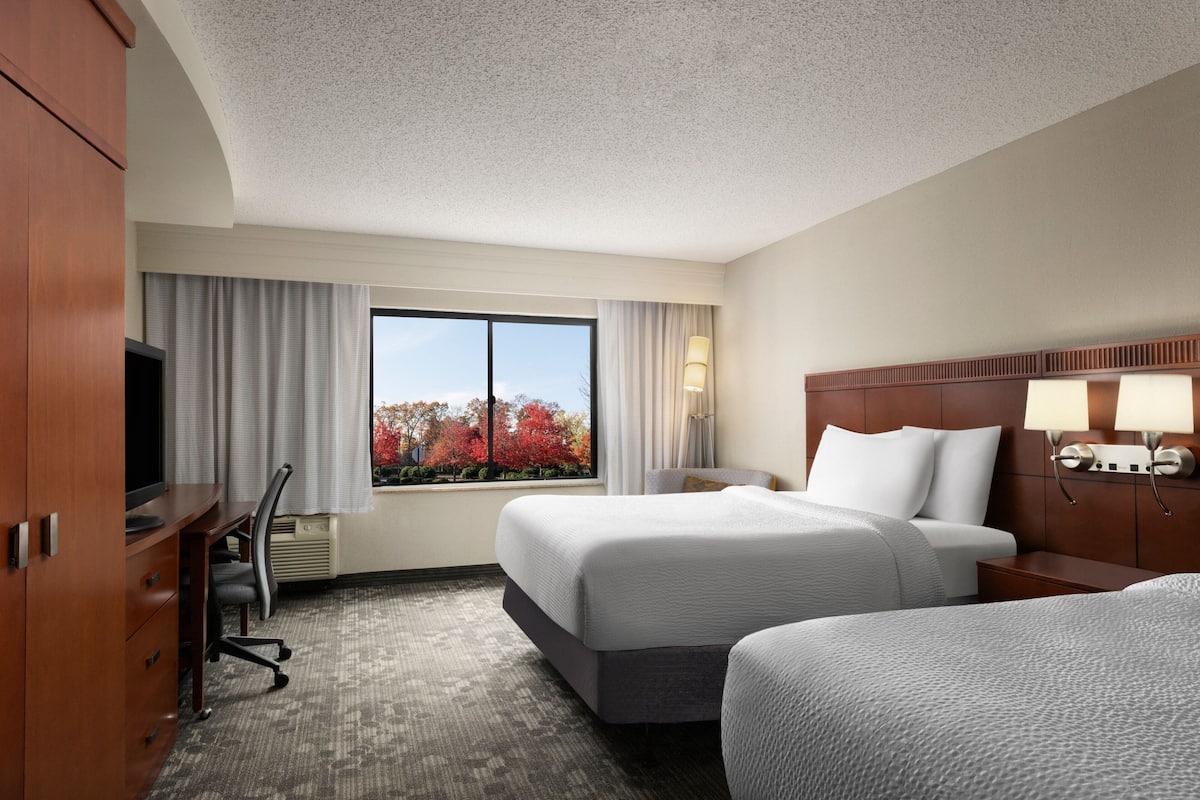 Une chambre d’hôtel avec un grand lit, un bureau avec une chaise, une télévision et une fenêtre donnant sur des arbres aux couleurs automnales.