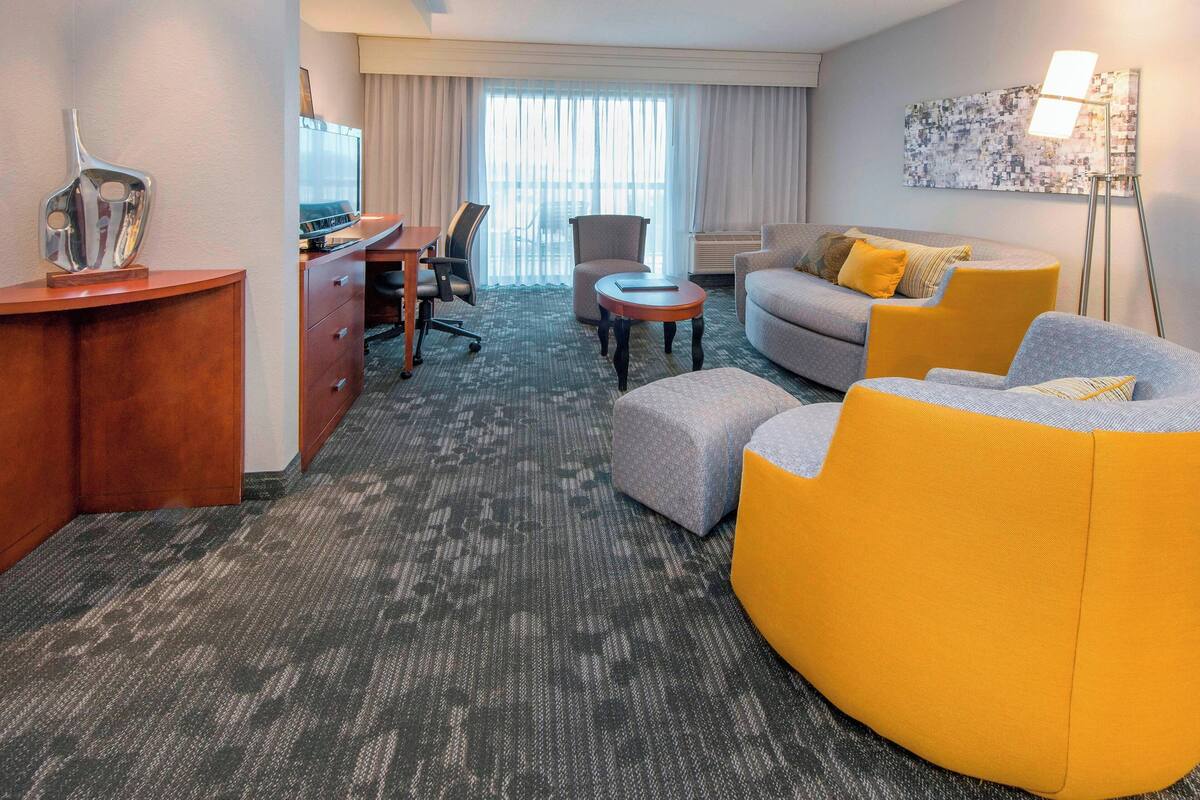 Ein Hotelzimmer mit Schreibtisch, grauem Sofa, gelbem Sessel, grauem Sessel, kleinem runden Tisch und Stehlampe.