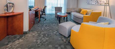 Una habitación de hotel con un escritorio, un sofá gris, un sillón amarillo, otro sillón gris, una mesita redonda pequeña y una lámpara de pie.