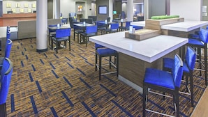 Un hall d’hôtel doté de plusieurs espaces de détente, notamment des coins aménagés comme des bars et des tables individuelles. On y trouve également un comptoir d’accueil et un grand écran en arrière-plan.