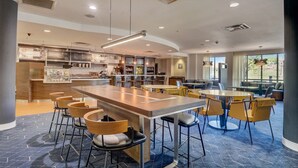 A modern restaurant with a central bar, high chairs, and a seating area with tables and chairs.