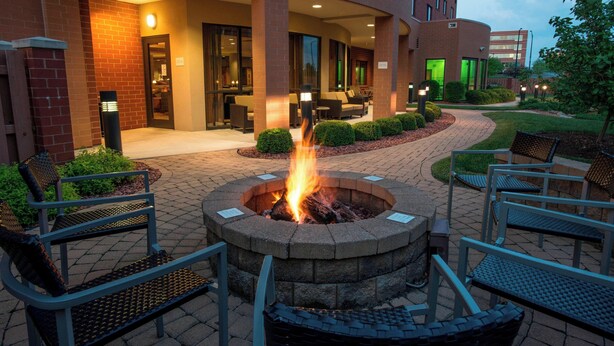 A fire pit surrounded by outdoor seating, a brick building with a covered patio, and a well-maintained garden.