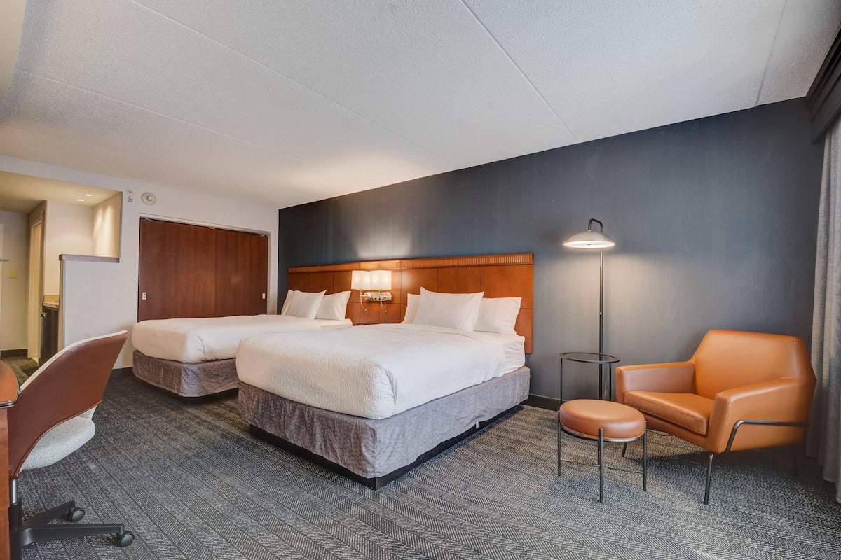 A hotel room with two beds, a desk, a chair, and a lamp.