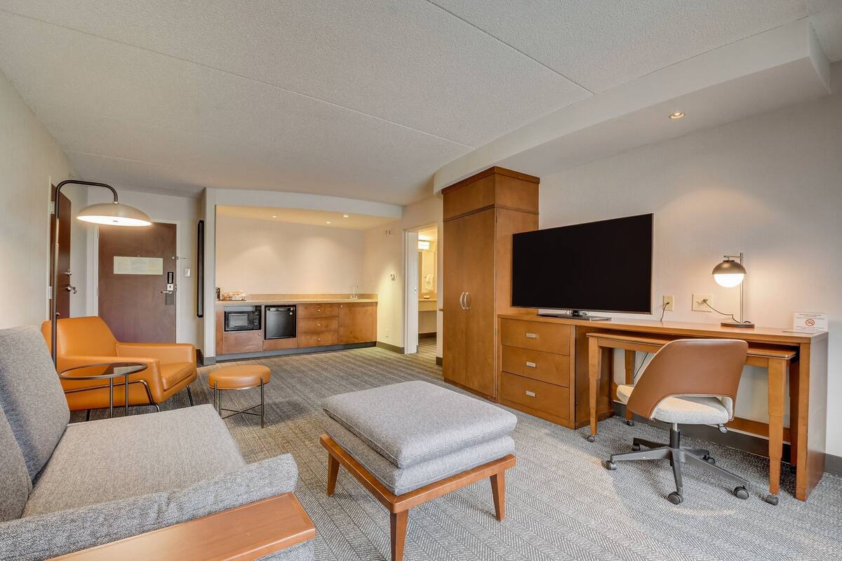 A hotel room with a sofa, ottoman, desk, chair, television, and kitchenette.