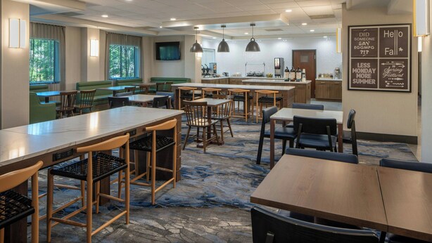 A modern dining area with multiple tables and chairs, a kitchen in the background, and a wall with motivational quotes. - Fairfield Inn & Suites by Marriott Lafayette South (Lafayette)