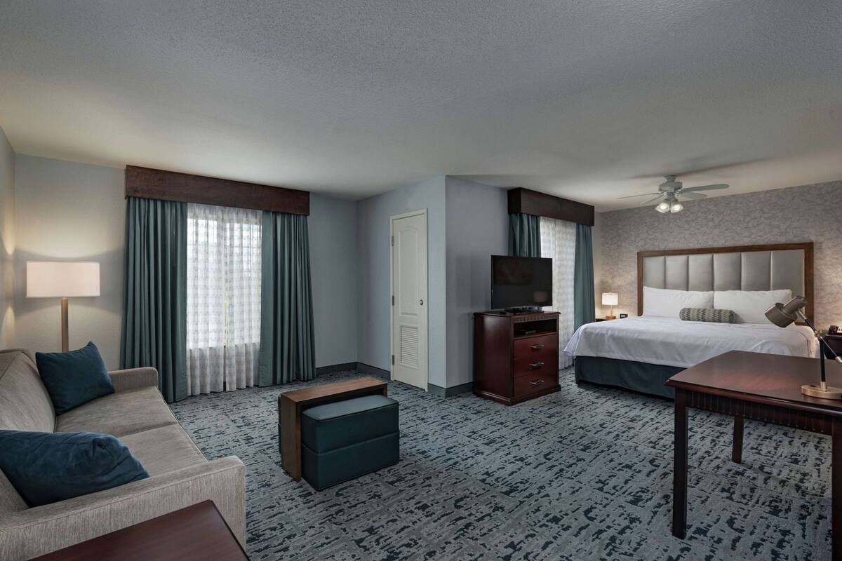 A hotel room with a bed, a TV, a desk, a chair, a sofa, and a window with curtains.