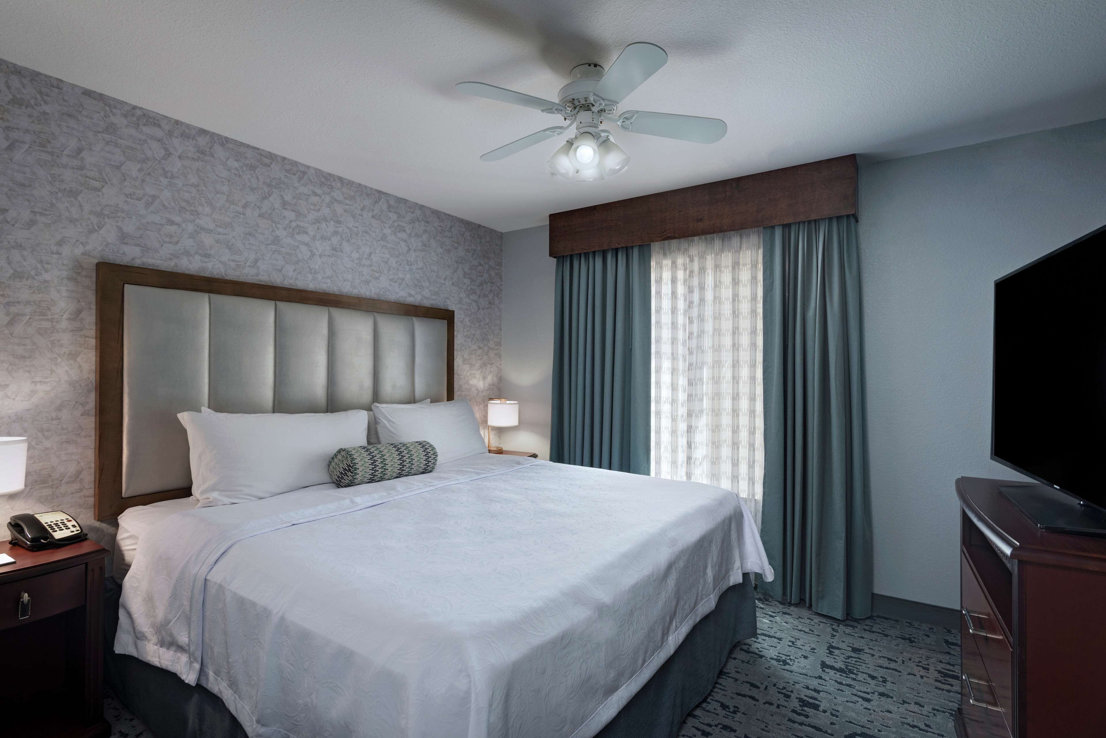 A hotel room with a large bed, a ceiling fan, a TV, and a nightstand with a phone.