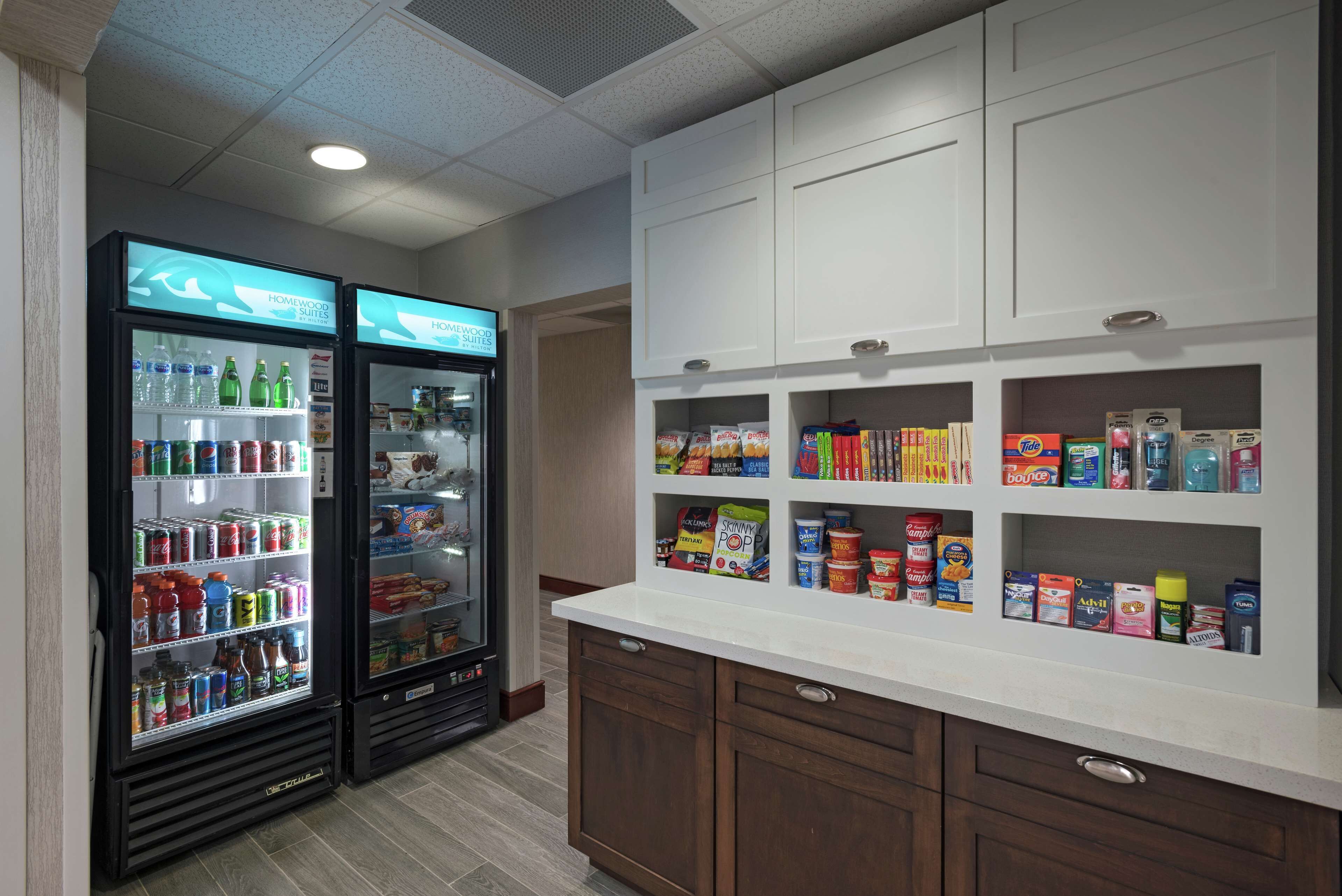 Una kitchenette con dos dispensadores de bebidas repletos de diversas opciones, una encimera blanca con estantes que contienen aperitivos y condimentos, y armarios de madera.