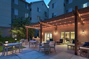 Un patio al aire libre con mesas y sillas, una pérgola con luces colgantes y la entrada a un edificio.