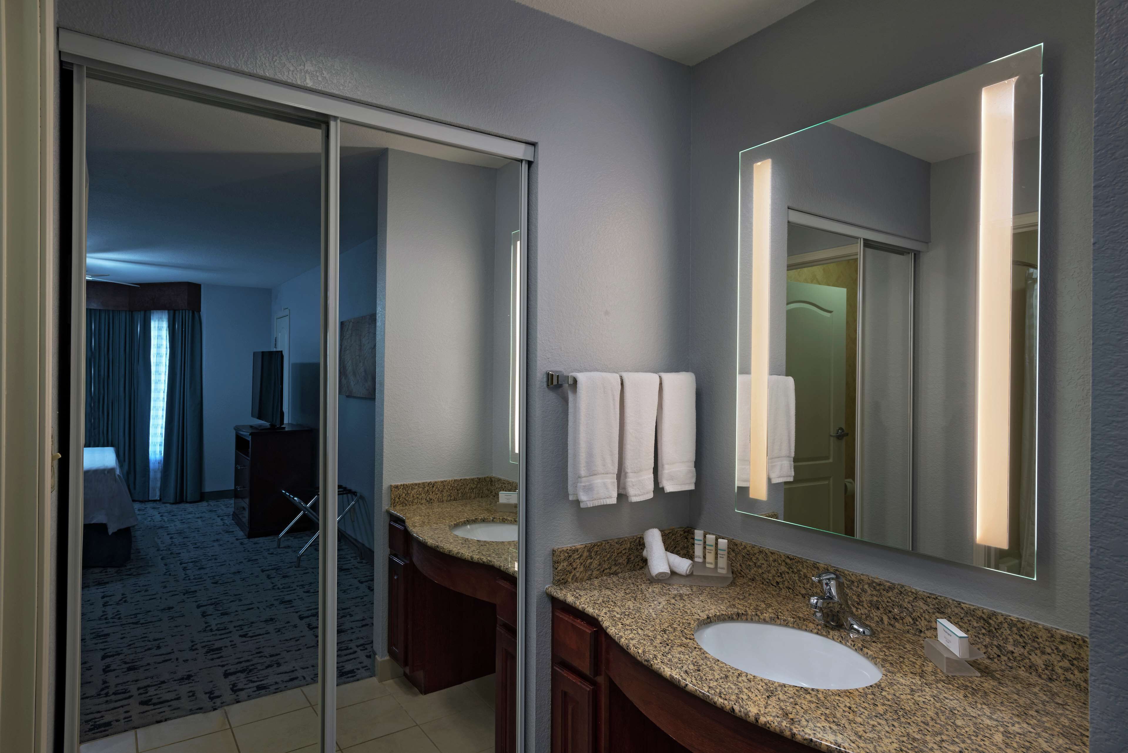 A modern bathroom with two sinks, a large mirror, and a sliding door leading to a bedroom.