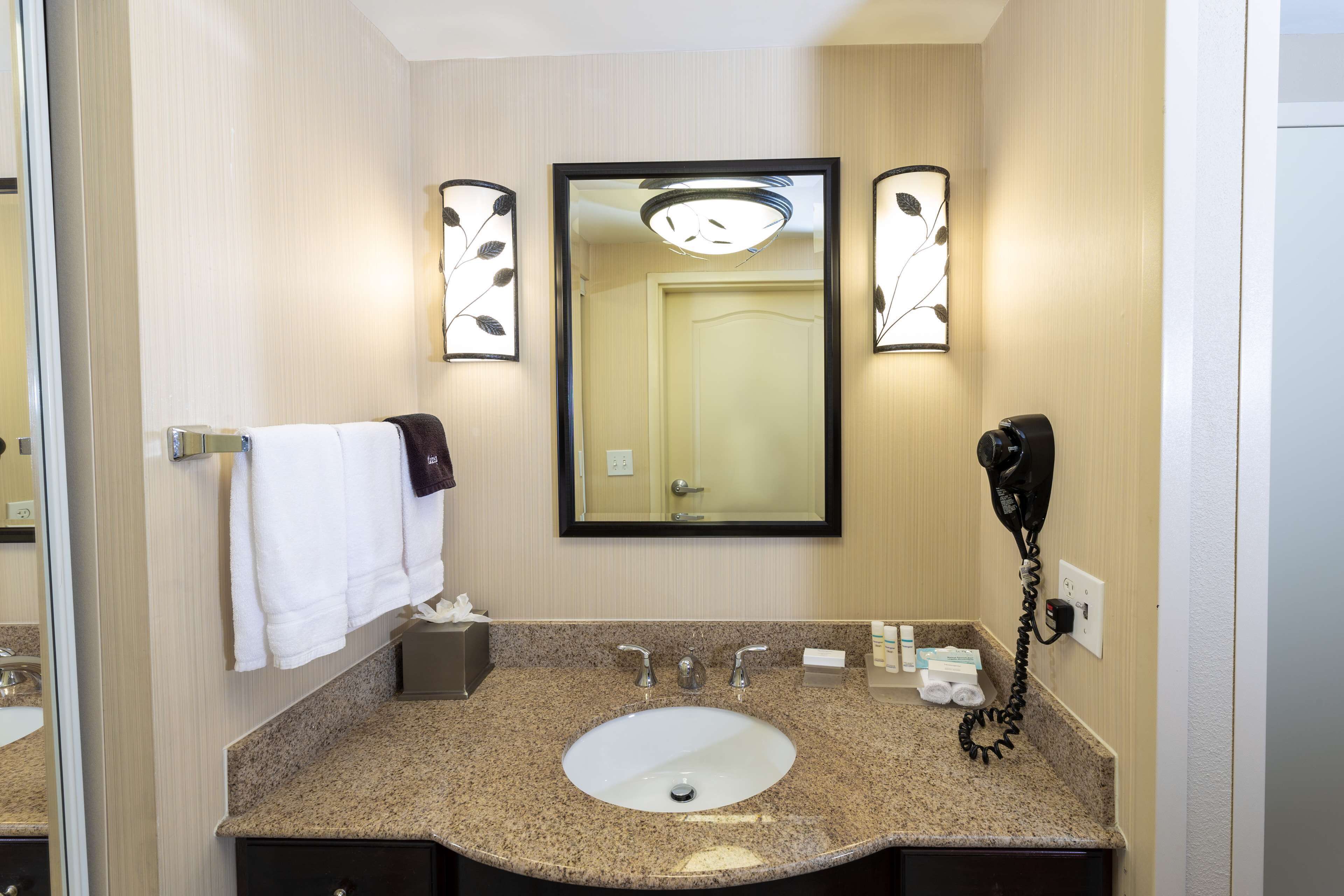 Une salle de bain d’hôtel avec un lavabo doté d’un plan de travail en granit, un miroir, un porte-serviettes, un téléphone et une petite boîte de mouchoirs.
