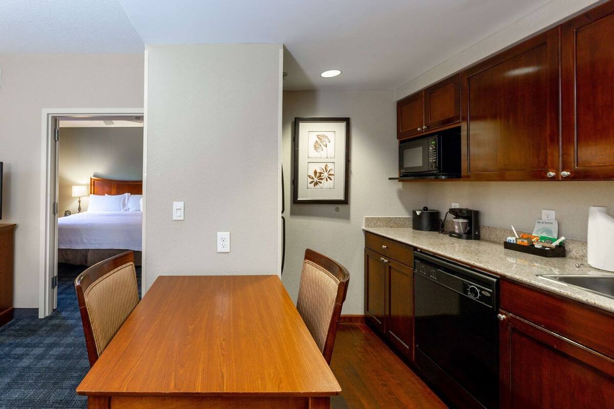 A hotel room with a wooden dining table, chairs, a kitchenette with a microwave, and a bed with white linens.
