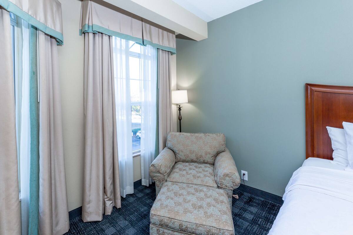 Une chambre d’hôtel avec un lit, une chaise, une lampe et une fenêtre avec des rideaux.