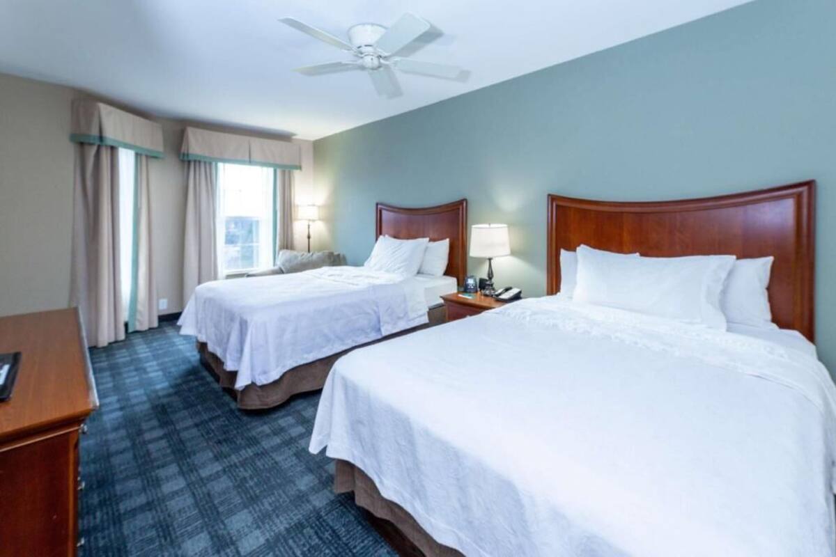 Une chambre d’hôtel avec deux lits, un bureau, une chaise, un ventilateur de plafond et une fenêtre avec des rideaux.