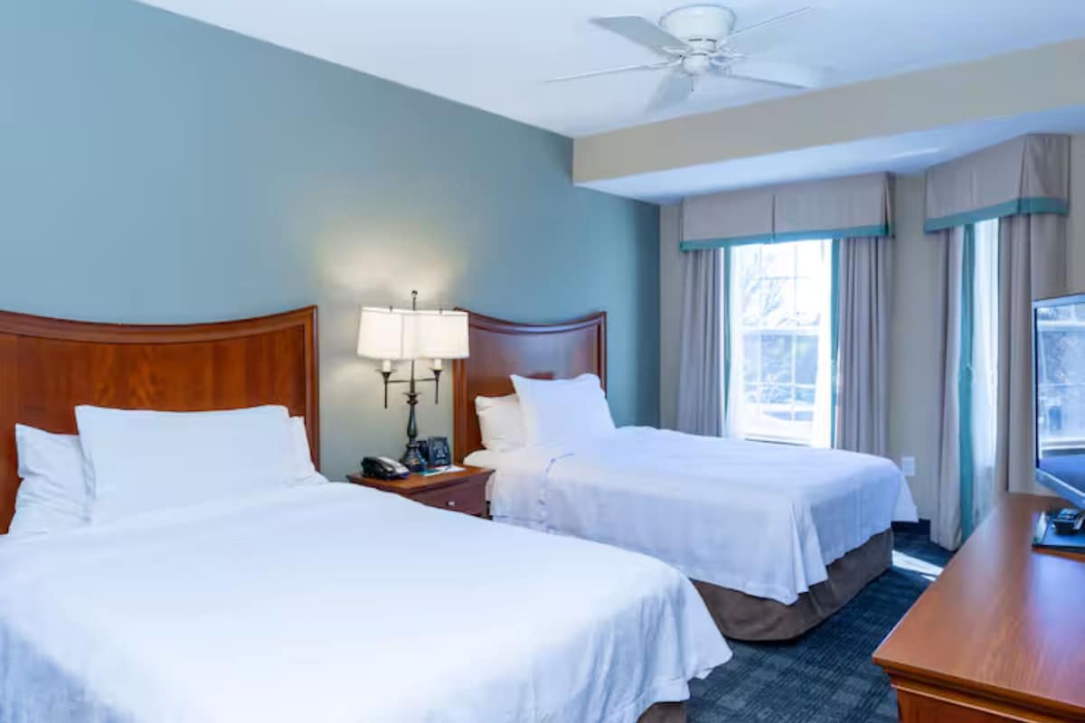 A hotel room with two beds, a desk, a television, a lamp, and a ceiling fan.