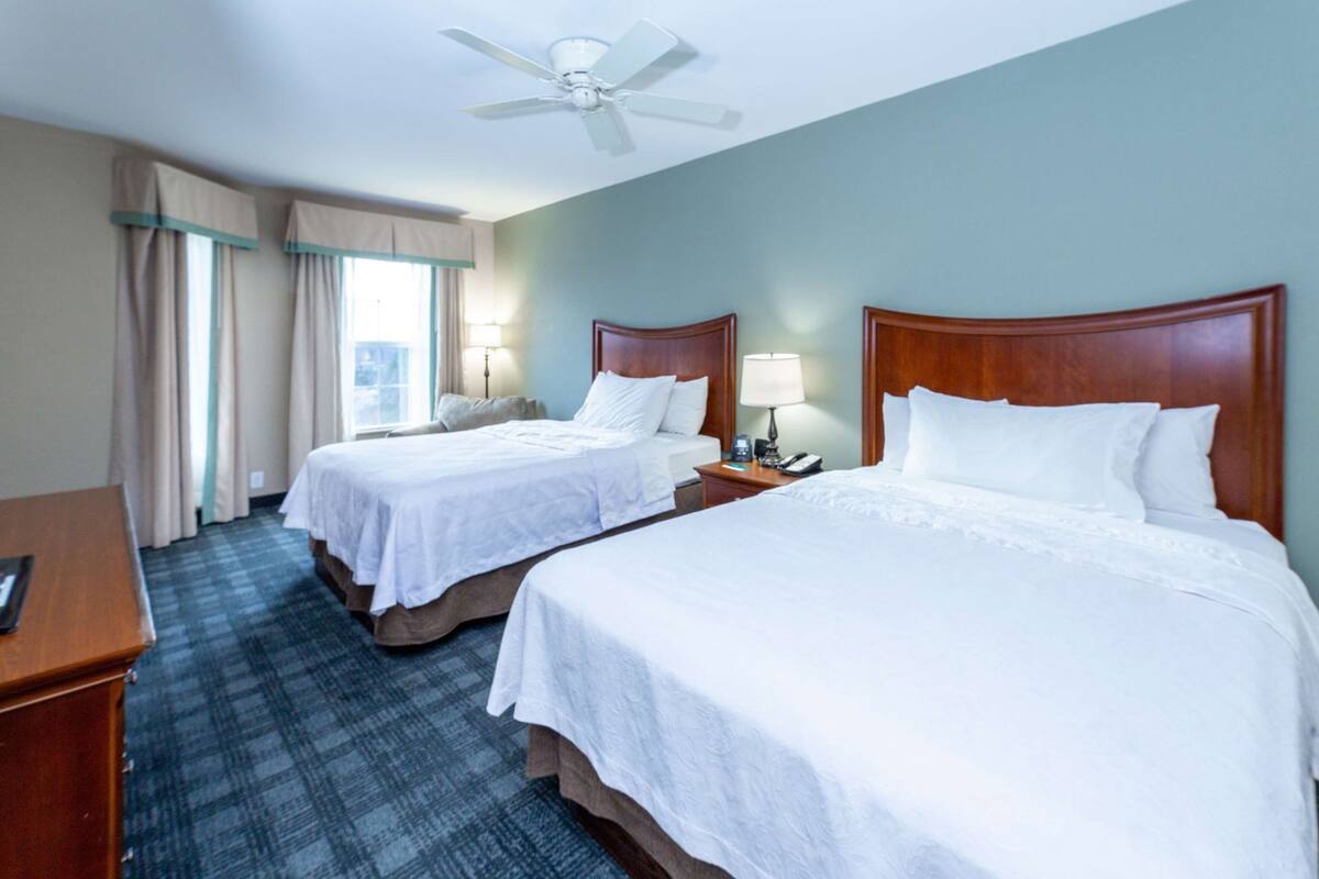 Une chambre d’hôtel avec deux lits, un bureau, une chaise, un ventilateur de plafond et une fenêtre avec des rideaux.
