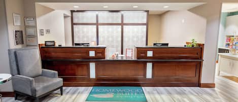 A reception area with a wooden desk, a grey armchair, and a doormat reading "Homewood Suites by Hilton".