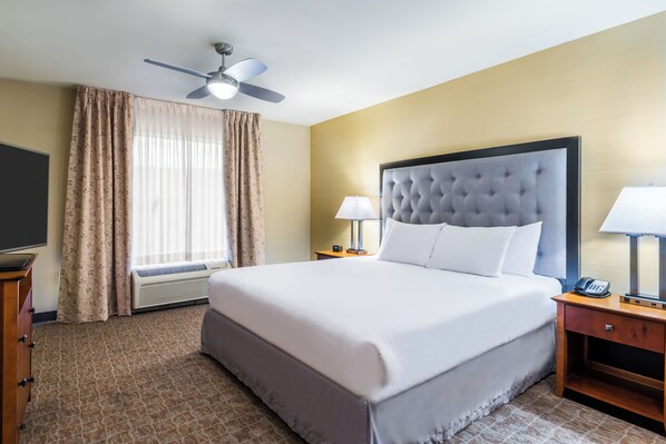 Una habitación de hotel con una cama grande, dos mesitas de noche con lámparas, un ventilador de techo y un ventanal con cortinas.