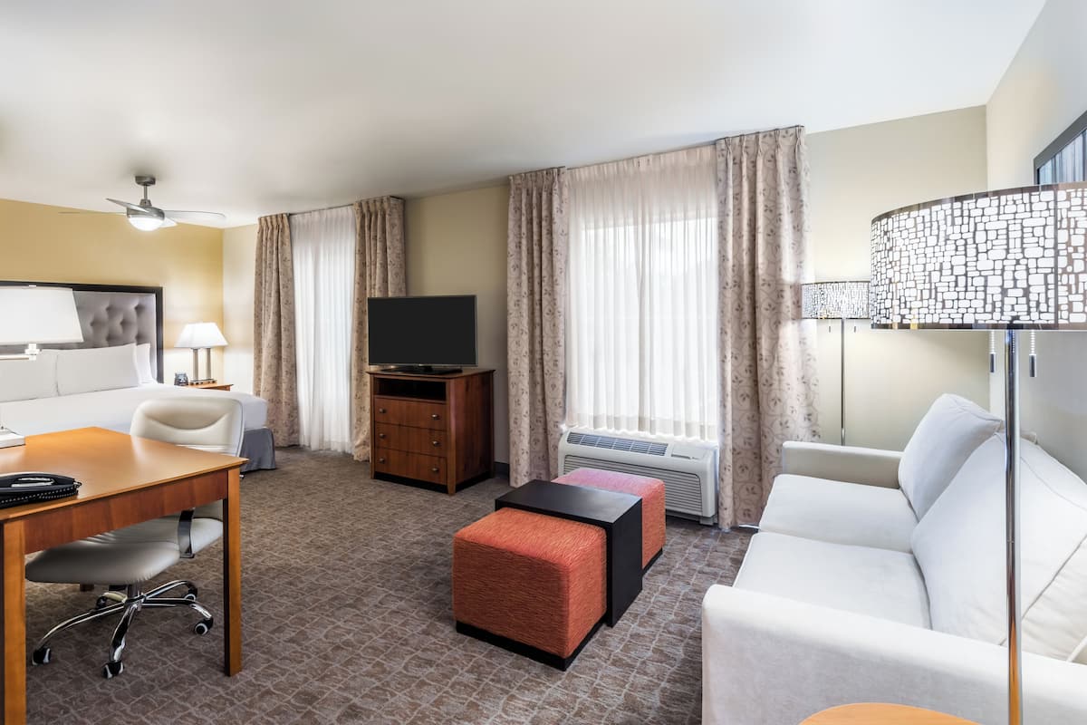 A hotel room with a bed, a desk with a chair, a television, a sofa, and a window with curtains.