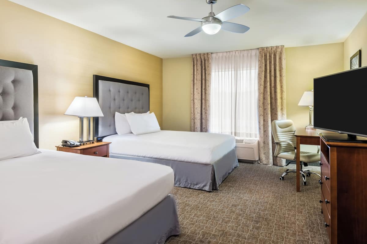 A hotel room with two beds, a desk, a chair, a television, and a ceiling fan.