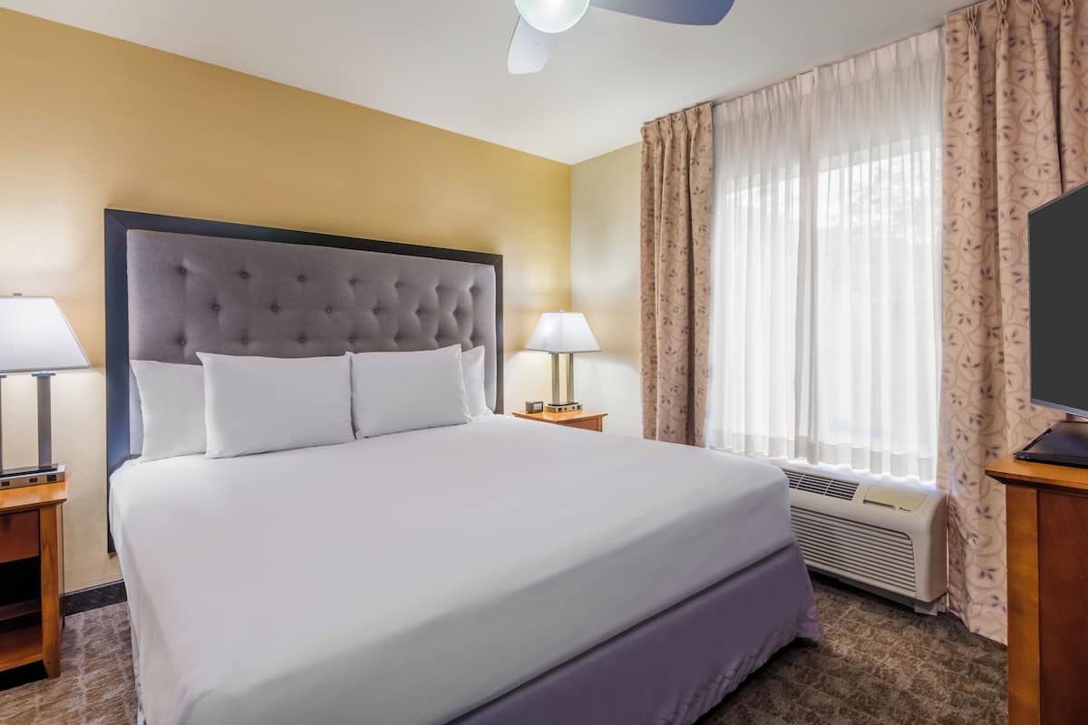 A hotel room with a large bed, two bedside tables, a lamp on each table, a TV on the right side, and a window with curtains.