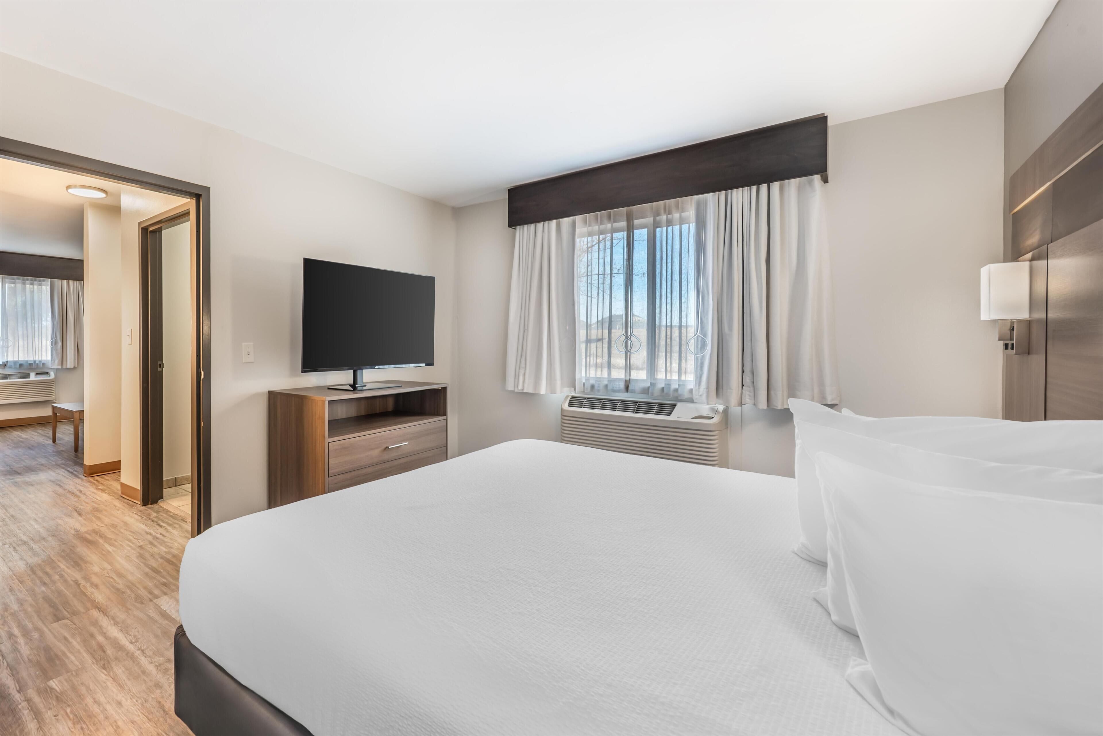 A hotel room with a large bed, a flat-screen TV, a desk with a chair, a window with curtains, and a wall-mounted lamp.