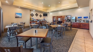 Une salle à manger d’hôtel avec plusieurs tables et chaises, un coin cuisine et un téléviseur fixé au mur.
