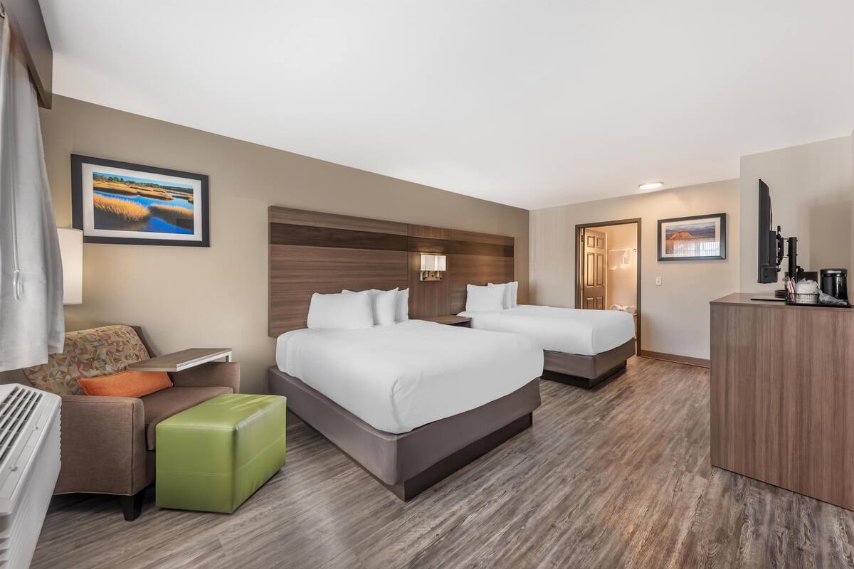 A hotel room with two beds, a green ottoman, a desk, and a TV.