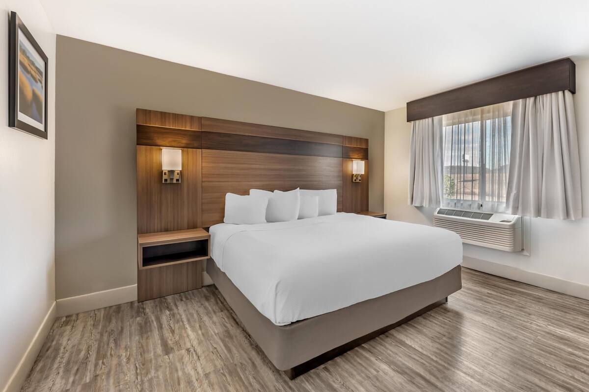 A hotel room with a large bed, wooden headboard, bedside lamps, a nightstand, and a window with curtains.
