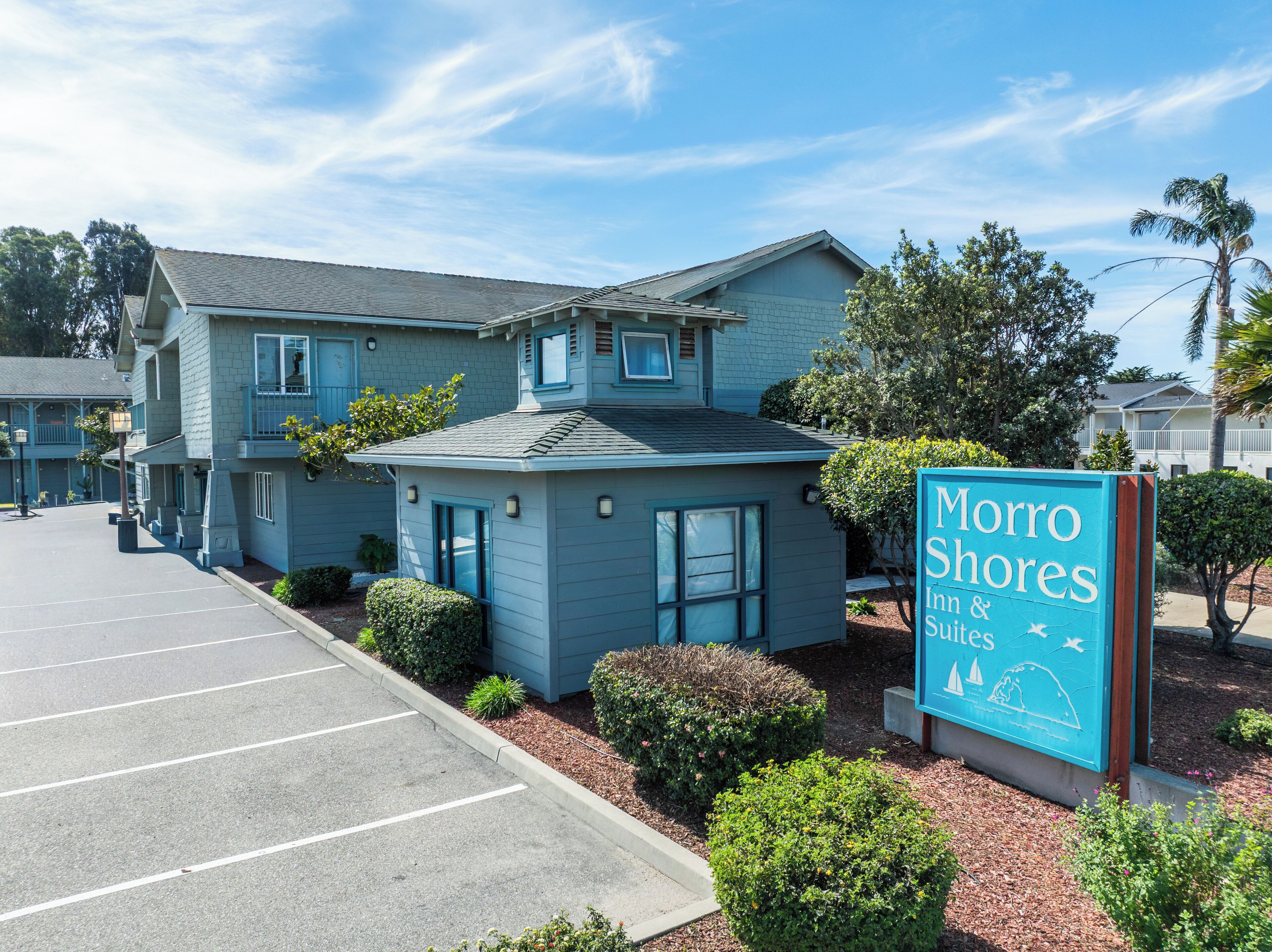 Un motel con un letrero que dice 'Morro Shores Inn & Suites' frente a un edificio de dos pisos con estacionamiento.