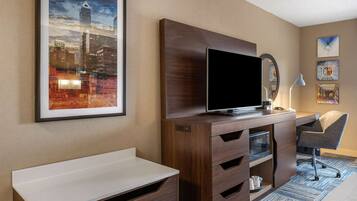 Habitación de hotel con un escritorio de madera, un televisor de pantalla plana, un espejo y una fotografía enmarcada de un paisaje urbano.