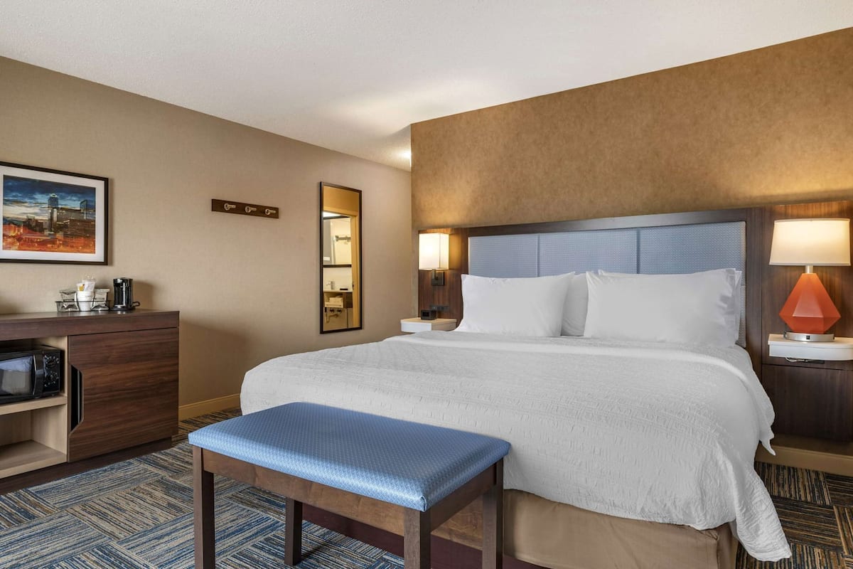 A hotel room with a bed, bedside tables, a desk with a microwave, a coffee maker, a mirror, and a picture on the wall.